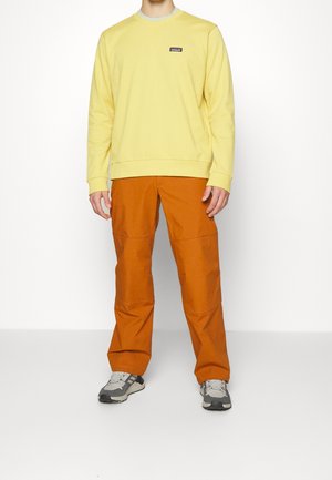 Persoon in een gele sweatshirt met lange mouwen, roest-oranje broek en grijze sneakers, staand tegen een witte achtergrond.