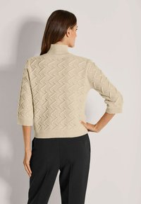 Frau mit langen dunklen Haaren, die einen beigen strukturierten Strickpullover mit Dreiviertelärmeln und schwarze Hosen trägt, steht mit der Hand in der Hüfte.