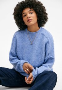 Hellblaues, gestricktes Sweatshirt mit weicher Textur, gerippten Bündchen und lässiger Passform. Legere Denim-Hosen und schlichte, goldene Schmuckakzente.
