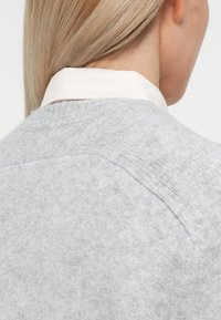 Pull en maille gris clair avec un col côtelé et des manches raglan, présentant une texture douce et un design minimaliste, porté par-dessus une chemise blanche à col.