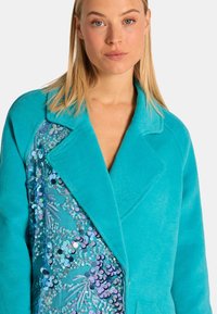 Türkise Fleece-Jacke mit einem glitzernden Paillettenmuster auf einer Seite, mit einem eingeknöpften Kragen und einer lockeren Passform.