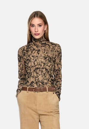 Woman wearing a beige turtleneck with black horse print, tan high-waist pants with brown belt, standing with hands in pockets against white background.