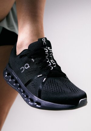 Chaussure de sport noire avec empeigne en maille ventilée, design distinctif de la semelle, logos de la marque blancs sur le côté et la languette, portée sur un pied levé.