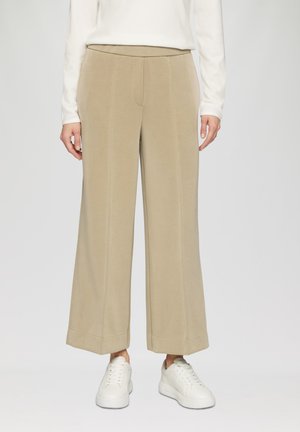 Pantalons large beige avec une texture lisse, présentant un design ajusté, des coutures verticales et un ourlet roulé, associés à des baskets blanches.
