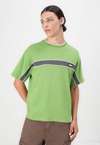 Green short-sleeve t-shirt in cotton, featuring a horizontal black stripe with white outline across the chest. Casual fit and rounded neckline.