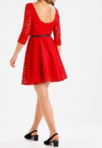 Robe rouge en dentelle avec un corsage ajusté et une jupe évasée, des manches trois-quarts, un dos ouvert et une ceinture noire à la taille. Sandales à talons noires.