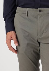 Lichtgrijze broek met een gestructureerde stof, voorzien van een ritssluiting, zijzakken en een enkele knoopsluiting. Vastgehouden door een hand tegen een witte achtergrond.