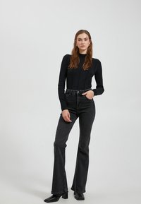 Zwarte lange mouwen top met verticale ribbels, gecombineerd met donker hoge taille jeans. Model draagt zwarte blokhak laarzen. Eenvoudige achtergrond.