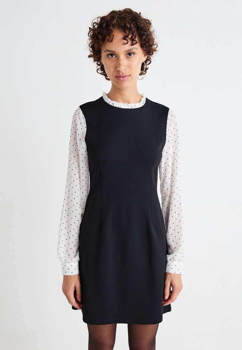 Mustat mekko, jossa on tiukka siluetti, pyöreä pääntie ja röyhelöreunus, yhdistettynä valkoiseen, pisaranmuotoiseen paitaan, jossa on pitkät hihat.