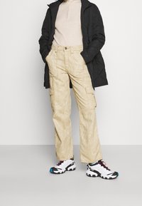 Beige cargobyxor med ett mönster av träådring, med flera fickor. Kombinerat med en svart jacka och vita sportskor. Slät textur.