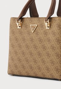 Brūna sintētiska soma ar strukturētu rakstu, zelta aparatūru un diviem rokturiem. Uz priekškara ir metāla logotipa trijstūris.