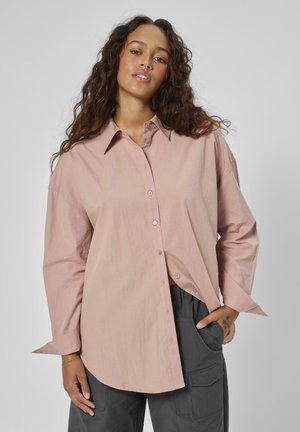 Pikkade lokkis juustega naine, kes kannab lahtist helekollast roosa nööpidega särki ja tumedaid halli värvi pükse, poseerib, hoides ühte kätt taskus.