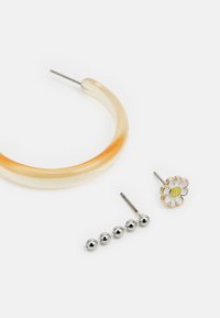 Três brincos: um argola translúcida amarelo-castanho, um par de brincos de bola prateada, e um brinco de flor dourada com um design em branco e verde.