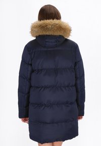 Mørkeblå pufferjakke med hætte kantet med pels, der har et quiltet design og lange ærmer. Stoffet ser skinnende og glat ud.