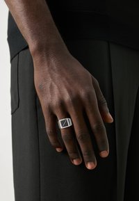 Sudraba zīmogs gredzens ar taisnstūra melnu akmeni, ar tekstūrētu joslu un ģeometrisku dizainu. Roka novietota uz tumša auduma.