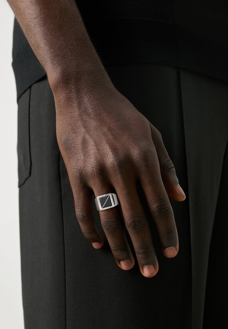 Sudraba zīmogs gredzens ar taisnstūra melnu akmeni, ar tekstūrētu joslu un ģeometrisku dizainu. Roka novietota uz tumša auduma.