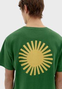Camiseta de algodón verde con un gráfico de sol amarillo en la parte posterior. Cuenta con mangas cortas y un diseño clásico de cuello redondo.