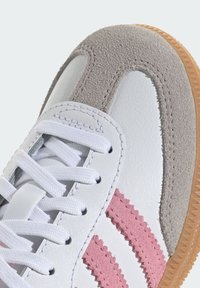 Biela kožená teniska s jemne sivými semišovými detailmi, tromi ružovými pruhmi a bielymi šnúrkami. Má textúrované povrchy a podrážku z gumy.