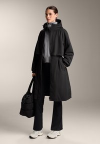 Fekete vízálló parka kapucnival és rugalmas derékpánttal, szürke zipzáros felsővel, fekete nadrággal és fehér sportcipővel párosítva, fekete táskát tartva.
