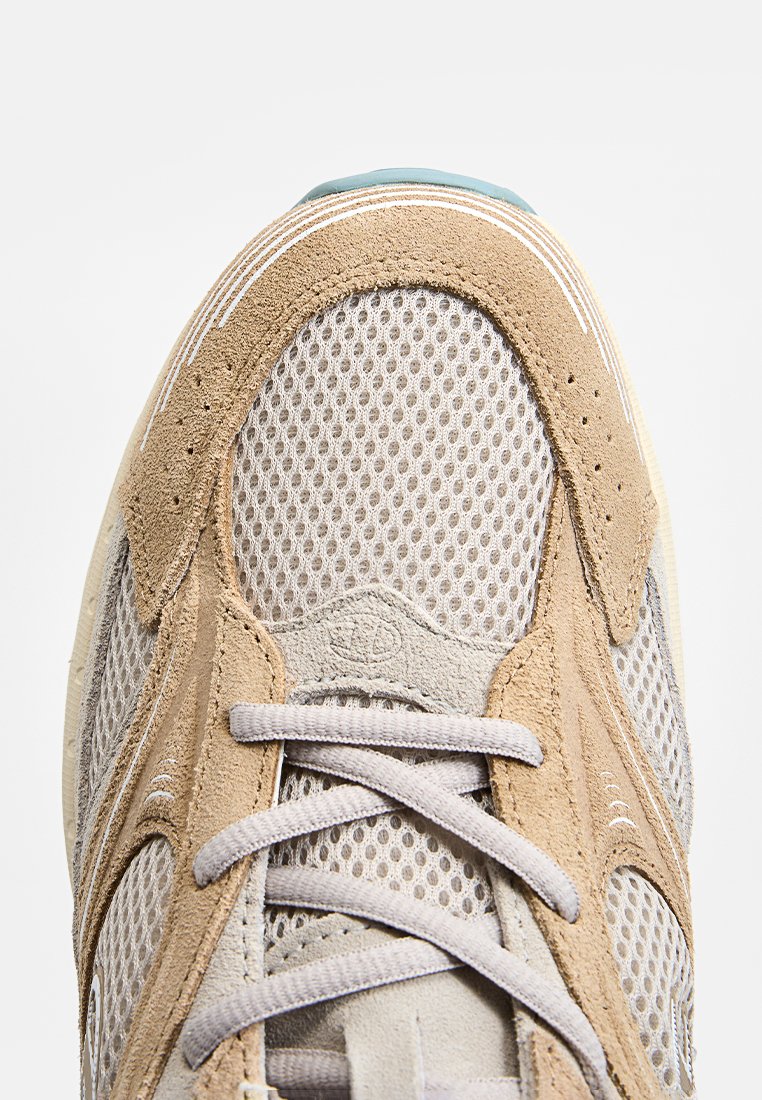 Beige und graue Sneaker mit Mesh-Einsätzen und Wildleder-Akzenten. Mit runder Zehenpartie und leichten Schnürsenkeln, ergänzt durch ein blaues Detail an der Sohle.