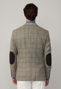 Blazer a quadri grigio con toppe in pelle marrone sui gomiti, vestibilità strutturata, chiusura a due bottoni e colletto blu a contrasto visibile sotto.