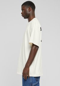 Crèmefärgad bomulls oversized t-shirt med korta ärmar, rund halsringning och en tryckt logotyp på baksidan. I kombination med blå denimjeans.