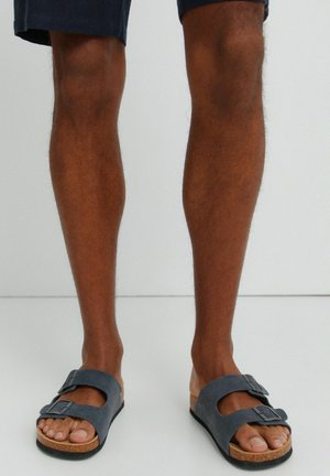 Gambe maschili che indossano pantaloncini blu navy e sandali blu scuro con due cinturini e fibbie su un pavimento bianco e sfondo semplice.