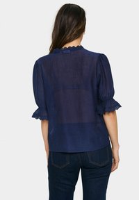 Blusa blu navy con maniche corte a sbuffo e bordo in pizzo scallop. Realizzata in un tessuto leggero e trasparente, presenta una superficie testurizzata.