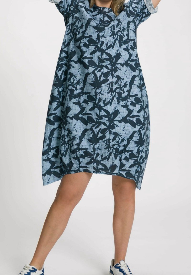 Femme portant une robe bleue ample avec des motifs abstraits foncés en forme de feuilles, debout les jambes écartées, chaussée de baskets blanches et bleues.