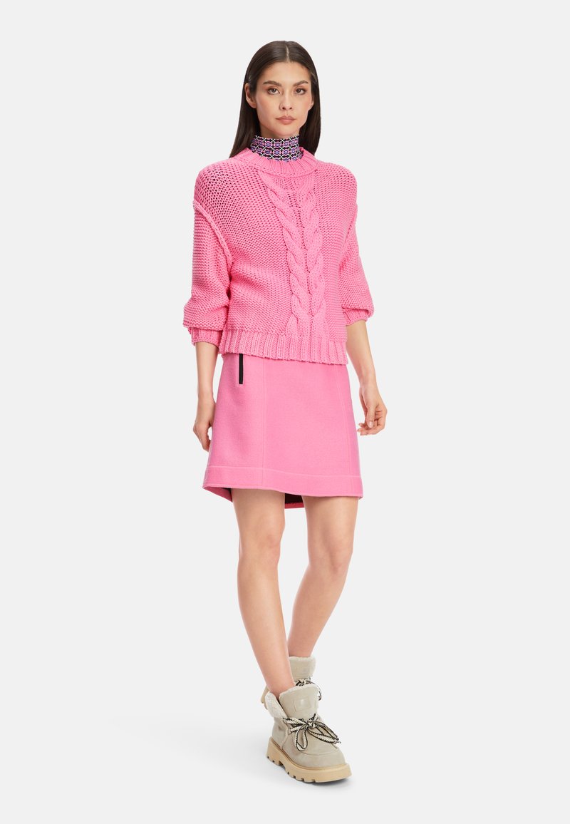 Heller pinker, grob gestrickter Pullover mit kurzen Ärmeln, kombiniert mit einem passenden pinken Minikörper. Beigefarbene Stiefel mit Kunstpelzbesatz und schwarzen Schnürsenkeln.