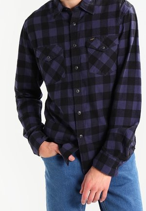 Mies, joka pukeutunut mustaan ja tummansiniseen ruudulliseen napilliseen paitaan, jossa on kaksi etutaskua, yhdistettynä sinisiin farkkuihin, seisoo käsi taskussa.