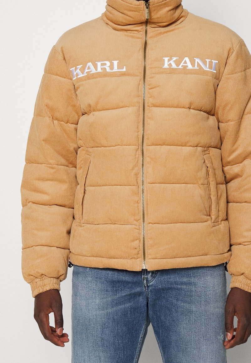 Uomo che indossa un piumino in corduroy beige con "KARL KANI" ricamato sul petto e jeans blu sbiaditi, in piedi con le braccia lungo i fianchi.