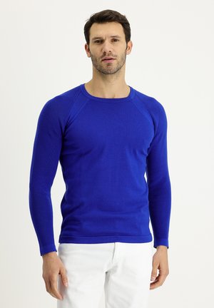 Maglione lavorato a maglia blu royal con texture a coste, maniche lunghe a raglan e scollatura a girocollo. Abbinato a jeans bianchi.