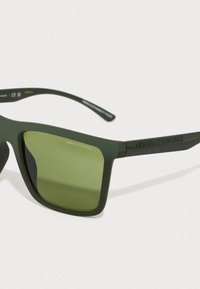 Žalios plastikinės saulės akinių su stačiakampiais lęšiais. Lęšiai yra žaliai tonuoti, o rėmelis turi iškilų užrašą "ARMANI EXCHANGE" ant šventyklų.