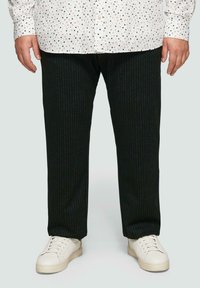Schwarze Nadelstreifenhose aus einem strukturierten Stoff, kombiniert mit weißen Sneakern und einem weißen Hemd mit einem gepunkteten Muster.