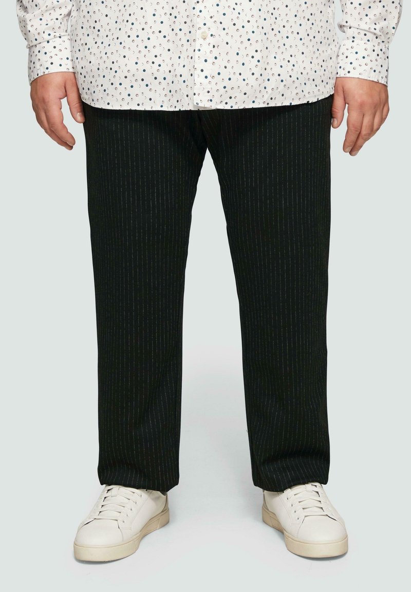 Schwarze Nadelstreifenhose aus einem strukturierten Stoff, kombiniert mit weißen Sneakern und einem weißen Hemd mit einem gepunkteten Muster.