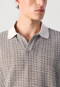 Young man wearing a beige and brown patterned knit polo shirt with a cream collar, facing slightly right against a plain background.