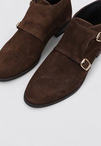 Zapatos de ante marrón con puntera puntiaguda, diseño de doble correa con detalles en hardware dorado y decoración de perforaciones.