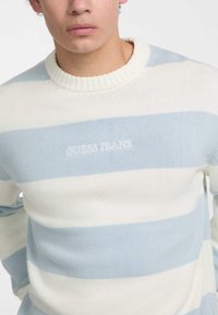 Svetlo modra in bela prugasta pletena pulover s rebrastim okrajem. Ima izvezen logotip "GUESS JEANS" v belem na prsih.