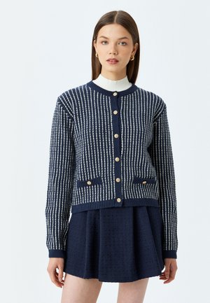 Cardigan marine avec des rayures blanches verticales, présentant des boutons dorés et deux poches avant. Poignets et ourlet côtelés, tissu tricoté texturé.