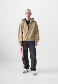 Beige Fleece-Zip-Hoodie, schwarze Cargo-Hosen, sportliche beige Sneakers und eine kleine rosa Tasche in der Hand, vor einem einfarbigen Hintergrund stehend.