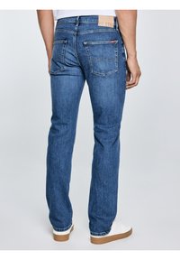 Vīrietis zilās taisna piegriezuma džinsa biksēs un baltos sporta apavos, stāvot pret vienkrāsainu gaišu fonu, skatoties no muguras.