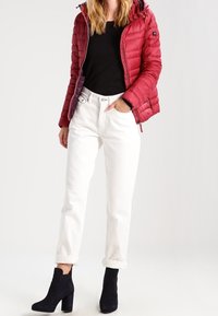 Rote Pufferjacke mit Kapuze, schwarzes Shirt darunter, weiße hochgekrempelte Hosen und schwarze Absatzstiefeletten. Glatte und strukturierte Materialien.