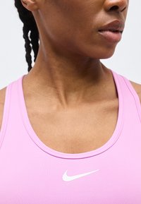 Κοντινό πλάνο ενός ατόμου που φορά ροζ αθλητικό μπλουζάκι Nike με πλεξούδες, δείχνοντας τον λαιμό και το κάτω μέρος του προσώπου γυρισμένα ελαφρώς προς τα πλάγια.