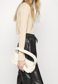 Vit flätad handväska med ett texturerat handtag, hålls av en person som bär en beige långärmad topp och svarta läderbyxor.