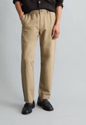 Mężczyzna w beżowych spodniach z elastycznym pasem i guzikami z przodu oraz czarnych mokasynach, stojący z jedną ręką w kieszeni na jednolitym tle.