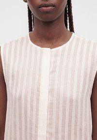 Personne aux cheveux foncés tressés portant un chemisier sans manches à rayures verticales beige clair et blanc avec un col rond.