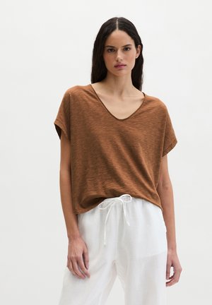 Frau trägt ein locker sitzendes braunes V-Ausschnitt-T-Shirt mit kurzen Ärmeln, das in weiße Hosen mit Kordelzug gesteckt ist, vor einem einfarbigen Hintergrund.