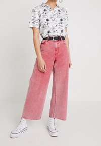 Lehká bílá košile s černým potiskem palmových stromů, spárovaná s vysokým pasem, širokými růžovými džínami a bílými teniskami na vyšší podrážce.