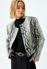Veste cintrée avec un motif de serpent en gris et noir, réalisée en tissu texturé, avec des boutons noirs.